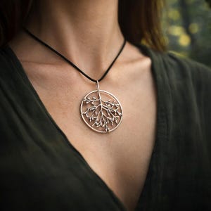 May include: Silver-toned pendant necklace featuring a circular design with a detailed tree branch motif. The pendant hangs from a black cord. The necklace is a nature-inspired accessory.