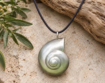 Puede incluir: Un colgante con forma de concha de nautilo en tono plateado suspendido de un cordón negro fino. El colgante presenta un acabado metálico liso con un diseño en espiral que se estrecha hacia el centro. La pieza está unida al cordón mediante una pequeña anilla metálica.