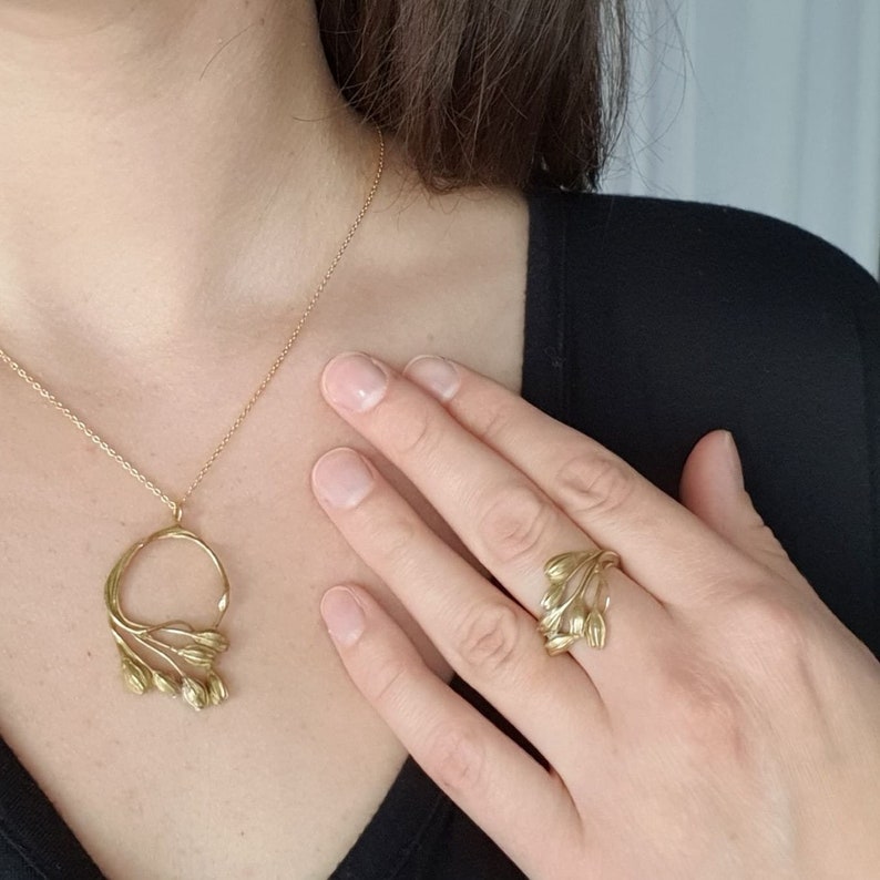 May include: Gold-toned jewelry set featuring a necklace with a pendant and a ring. The pendant has a circular design with leaf-like details. The ring also has a leaf-like design. The jewelry is worn on a person wearing a black top.