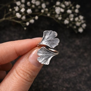 Op de afbeelding: Een zilverkleurige ring met een dubbel ginkgo blad ontwerp. De bladeren hebben gedetailleerde nerven en een getextureerd oppervlak. De ring wordt getoond tegen een donkere achtergrond met witte bloemen.