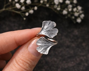 Puede incluir: Un anillo de tono plateado con dos motivos de hojas de ginkgo. El metal tiene un acabado oxidado y texturizado que resalta las venas de las hojas. El diseño es abierto, con las dos hojas orientadas en direcciones opuestas en la banda.