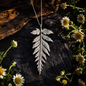 May include: A silver-toned necklace featuring a detailed, textured fern leaf pendant suspended from a delicate chain. The metallic leaf displays intricate veining and serrated edges, capturing a realistic botanical form in a polished finish.