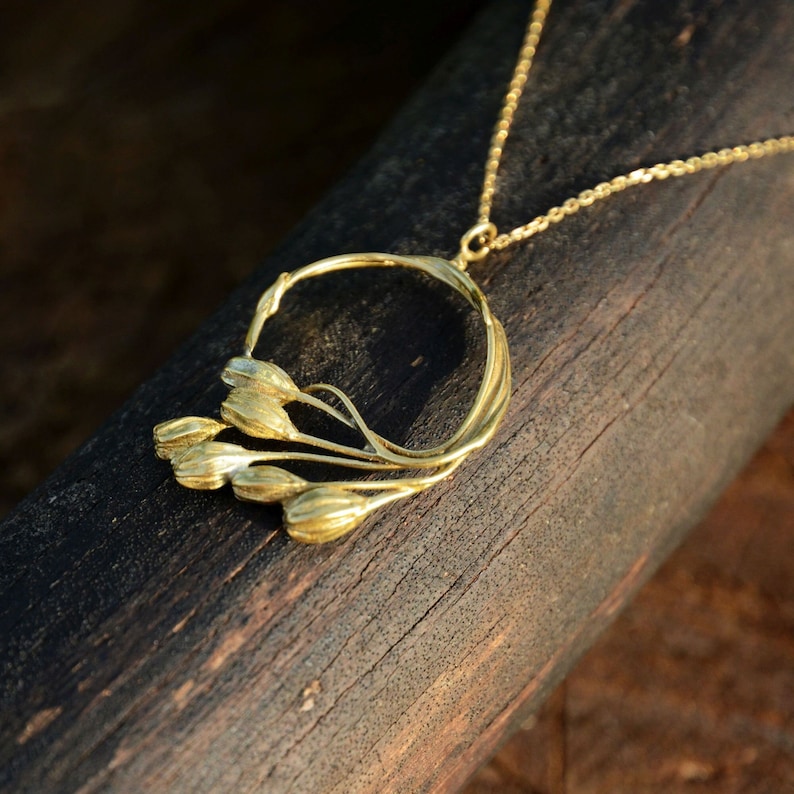 May include: A gold-colored necklace featuring a floral design. The pendant has a circular frame with stylized flower buds and stems. The chain is delicate, and the necklace is displayed on a dark wooden surface.