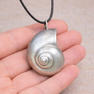 Puede incluir: Un colgante plateado con forma de concha de nautilus, suspendido de un cordón negro. La concha presenta un diseño en espiral detallado. La joya se sostiene en una mano sobre un fondo neutro. Es un collar.
