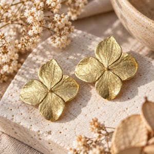 May include: A pair of gold-toned stud earrings shaped like four-petaled flowers. Each earring features a textured, metallic surface with visible veins radiating from the center, mimicking the appearance of a natural hydrangea blossom.