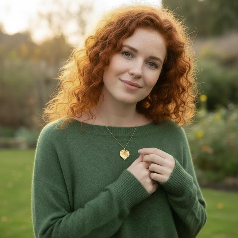 Puede incluir: Un collar con colgante de hoja dorada. El collar se lleva sobre un su&eacute;ter verde. El colgante de hoja est&aacute; detallado con venas y cuelga de una delicada cadena. El fondo es una escena borrosa al aire libre.