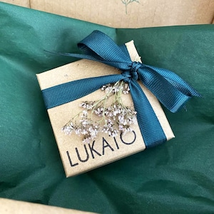 May include: A small, square, kraft paper gift box tied with a teal ribbon and a bow. The box is adorned with dried flowers and the word "LUKAITO" is printed on the front. The box rests on a bed of green tissue paper.