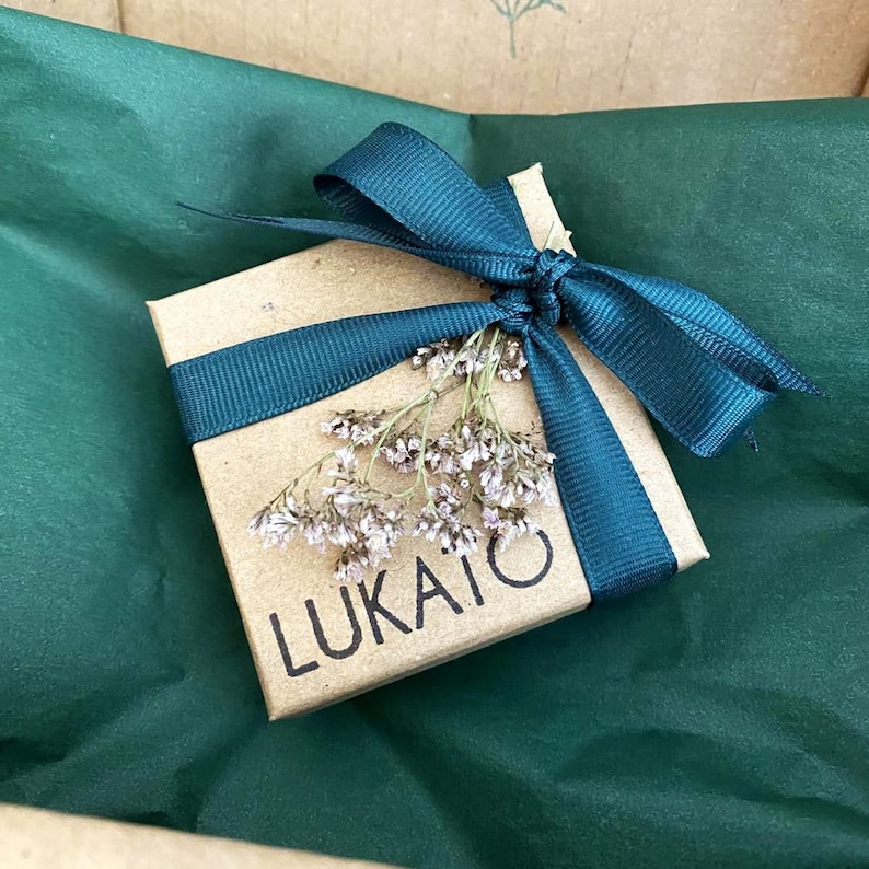 May include: A small, square, kraft paper gift box tied with a teal ribbon and a bow. The box is adorned with dried flowers and the word "LUKAITO" is printed on the front. The box rests on a bed of green tissue paper.