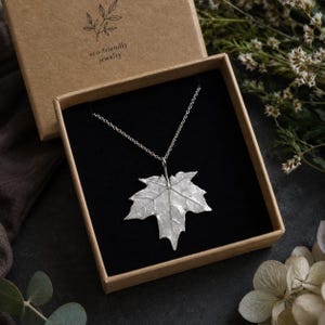 May include: A silver-colored leaf pendant necklace displayed in a brown box with the text "eco-friendly jewelry". The necklace features a detailed maple leaf design on a delicate chain, set against a black interior.