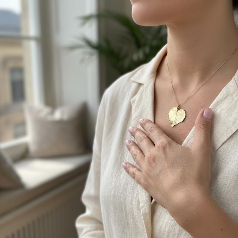 Puede incluir: Collar con colgante de hoja de color dorado. El colgante es una r&eacute;plica detallada de una hoja, suspendida de una delicada cadena. El collar se lleva sobre una camisa clara con cuello.