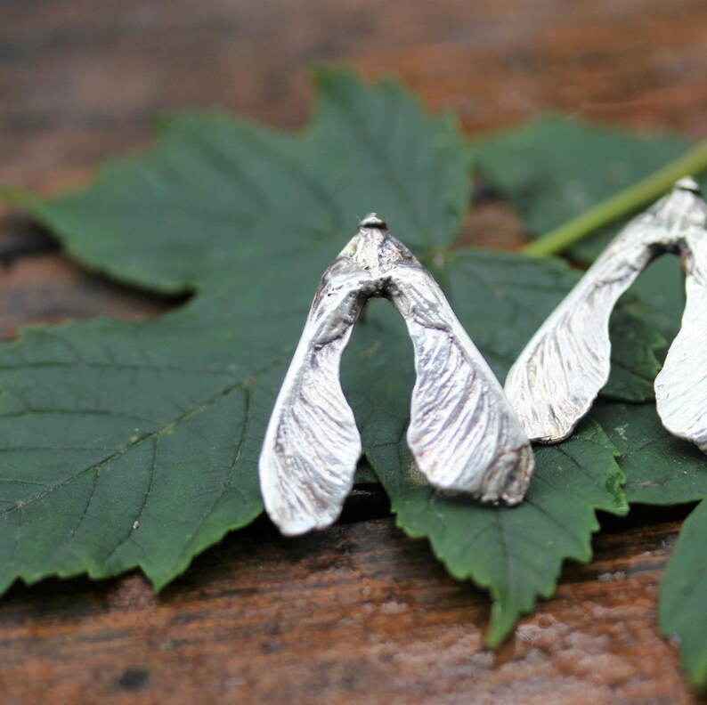 May include: A pair of silver-toned earrings shaped like maple tree samara seeds. Each earring features a textured, metallic surface that mimics the veined appearance of a winged seed pod, with a pointed top and two distinct, elongated lobes.