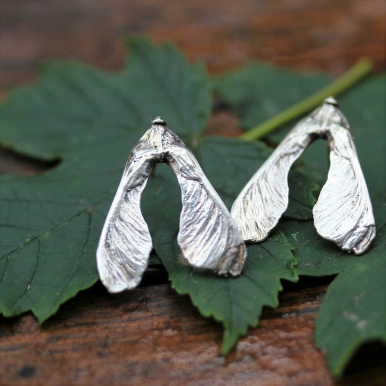May include: A pair of silver stud earrings shaped like maple tree samara seeds. Each earring features a textured, metallic surface that mimics the organic, veined appearance of a winged seed pod, with a pointed top and two distinct, flared lobes.