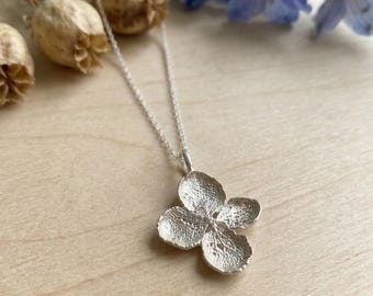 Puede incluir: Un collar con colgante plateado con un diseño de flor de cuatro pétalos. El colgante tiene una superficie texturizada y cuelga de una delicada cadena de plata. El fondo incluye vainas de semillas secas y flores azules.