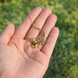 May include: A gold-colored necklace featuring a circular pendant with a floral design. The pendant has a ring shape with delicate, bud-like elements. The necklace is displayed against a blurred green background.