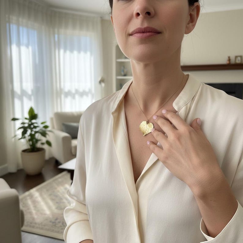 Puede incluir: Un collar con colgante de hoja dorada. El colgante, una detallada representaci&oacute;n de una hoja, cuelga de una fina cadena de oro. La mujer lleva una blusa color crema.