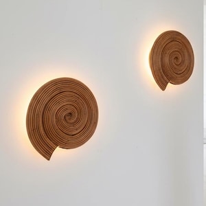 Op de afbeelding: Twee wandlampen in spiraalvorm, die op schelpen lijken. Gemaakt van geweven, lichtbruin materiaal, stralen ze een warm licht uit. Het ontwerp voegt een natuurlijke, maritieme esthetiek toe aan de ruimte.