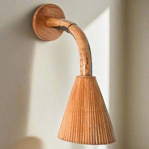 May include: A wall-mounted light fitting with a curved wooden arm and a conical woven shade. The shade is made of natural materials and has a warm, rustic look.