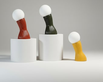 May include: Three modern table lamps with a white spherical bulb and a textured, curved base. The bases are in shades of red, green, and yellow, each set on a white cylindrical pedestal. The lamps have a minimalist design.