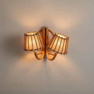 Puede incluir: Aplique de pared doble con dos pantallas. Las pantallas son cilíndricas, hechas de un material tejido de color claro y con un borde decorativo. La lámpara está hecha de un material marrón claro, posiblemente bambú o ratán.