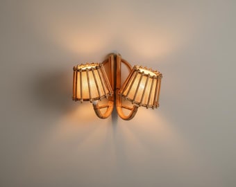 May include: A wall-mounted light fixture with two lampshades. The shades are cylindrical, made of a light-colored woven material, and trimmed with a decorative edge. The fixture is made of a light brown material, possibly bamboo or rattan.