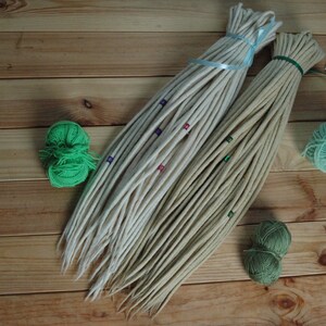 May include: Two bundles of white and beige wool dreadlocks with colorful beads. The dreadlocks are tied together with blue ribbon. Two balls of green yarn are on the wooden surface.