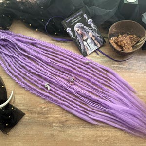 May include: A bundle of lavender dreadlocks with a lighter purple ombre effect. The dreadlocks are adorned with small silver charms. A book titled "Dreads Forest" and a decorative bowl are also visible.