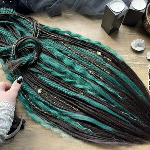 May include: A set of synthetic hair extensions featuring a mix of dark brown and teal green strands. The hair includes a combination of straight, wavy, and braided textures, accented with thin metallic gold threading and small decorative beads.