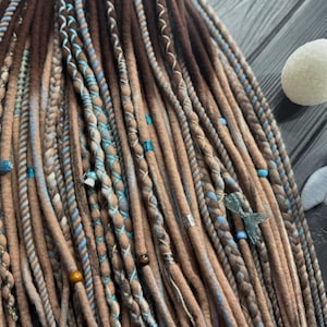 Puede incluir: Primer plano de rastas marrones, beige y azules. Las rastas están adornadas con cuentas azules y detalles decorativos. También se ve una vela blanca en forma de huevo y una figura de ballena azul.
