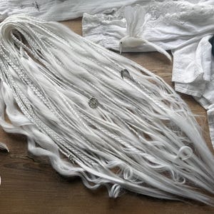 Puede incluir: Dreadlocks blancos y plateados con secciones trenzadas y sueltas, adornados con abalorios plateados. Las extensiones de cabello están dispuestas sobre una superficie de madera con tela blanca, pájaros decorativos y una vela.
