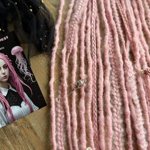 Puede incluir: Se muestran rastas rosas con varias texturas. Es visible una tarjeta con el texto "Dread5 Forest" y una ilustración de una mujer con cabello rosado. También hay varios accesorios decorativos para el cabello.