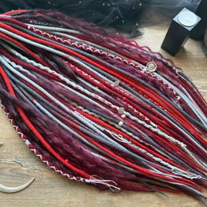 May include: A collection of burgundy, red, and gray dreadlocks, some with white stitching and decorative beads. The dreadlocks are arranged on a wooden surface, with a black comb and a silver crescent moon-shaped hair accessory.
