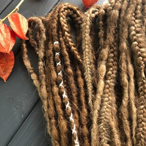a collection of braided hair strands in various colors and textures, including brown, blonde, and red, arranged on a wooden surface. There are also some dried orange flowers in the background.