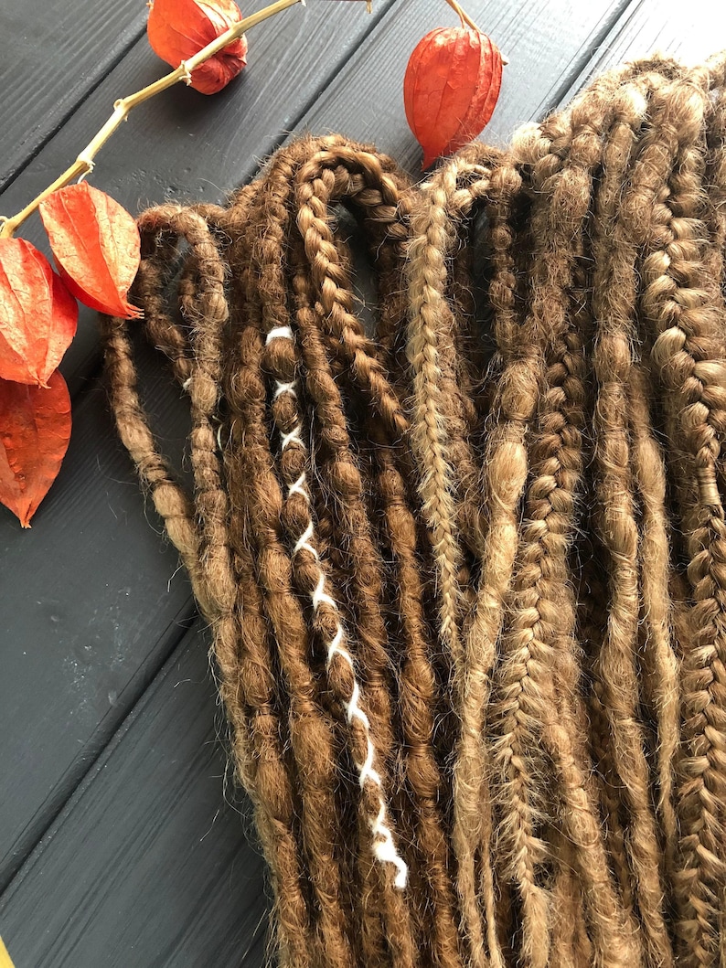 a collection of braided hair strands in various colors and textures, including brown, blonde, and red, arranged on a wooden surface. There are also some dried orange flowers in the background.