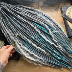May include: A set of gray, white, and teal dreadlocks with decorative charms. The dreadlocks are arranged on a wooden surface, with a book and comb visible in the background. The Dreads Forest logo is in the upper left corner.