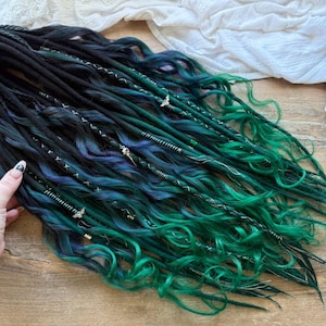 May include: A bundle of synthetic dreadlocks in shades of black, teal, and blue. The dreads are styled with decorative silver accents and are arranged on a wooden surface. The logo "Dreads Forest" is visible in the background.