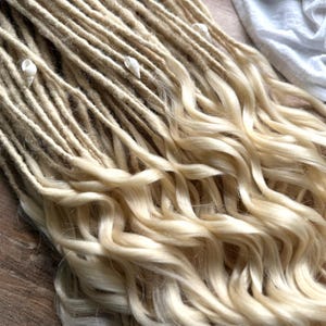 Peut inclure: Dreadlocks blonds avec des extrémités ondulées, ornés de petits accents de coquillages blancs. Les dreads passent d'un blond plus foncé à un blond plus clair et ondulé. Le logo Dread5 Forest est visible en bas à gauche.