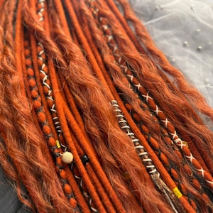 Può includere: Primo piano di dreadlock arancioni con trecce decorative. Alcune trecce hanno motivi a punto croce bianchi, perline nere e perline di legno. Il logo Dread5 Forest è visibile in alto a destra. Una piccola zucca è in basso a sinistra.