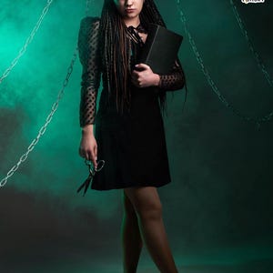 Portrait of a model wearing dark green and black synthetic dreadlocks and fishtail braids. She is looking at the camera, holding a black skull candle holder. The image highlights the natural blend of the dreads with her dark hair and fringe.