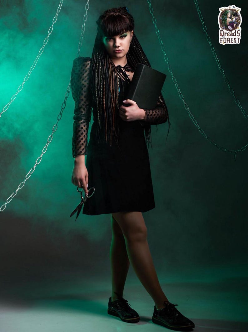 Portrait of a model wearing dark green and black synthetic dreadlocks and fishtail braids. She is looking at the camera, holding a black skull candle holder. The image highlights the natural blend of the dreads with her dark hair and fringe.