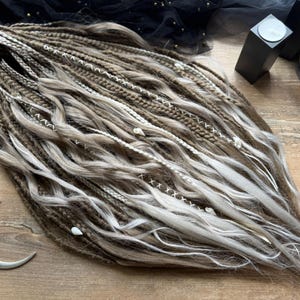 May include: Handmade dreadlocks in brown, grey, and white. The dreads are styled with white thread and small decorative elements. The image is taken from above, showing the texture and colour variations. A decorative crescent moon is visible.