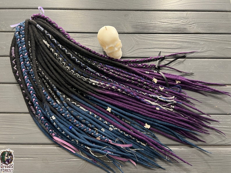 Puede incluir: Un conjunto de rastas sint&eacute;ticas con hebras de lana negra, azul oscuro y p&uacute;rpura. Las rastas est&aacute;n dispuestas en forma de abanico sobre una superficie de madera.