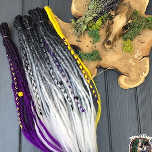 May include: A set of synthetic dreadlocks with ombre coloring, transitioning from black to gray to white. The dreadlocks are accented with purple and yellow yarn, and a yellow bead is visible on one of the purple strands.