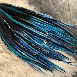 May include: A set of blue and black synthetic dreadlocks with silver beads and charms. The dreadlocks are arranged in a fan shape and are lying on a white fluffy surface.