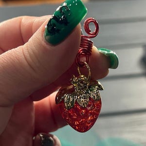 A hand holding a red wire charm with a green leaf design and a red strawberry charm.