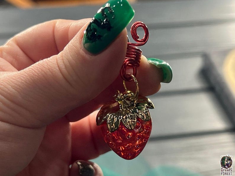 A hand holding a red wire charm with a green leaf design and a red strawberry charm.
