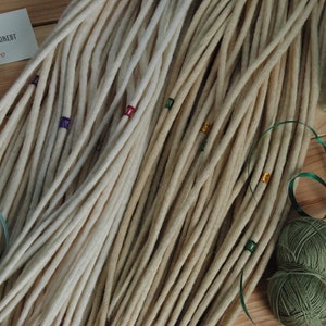 May include: A bundle of off-white wool dreadlocks with colorful beads. The dreadlocks are arranged in a fan shape on a wooden surface. There are two balls of yarn in the background, one green and one dark green.