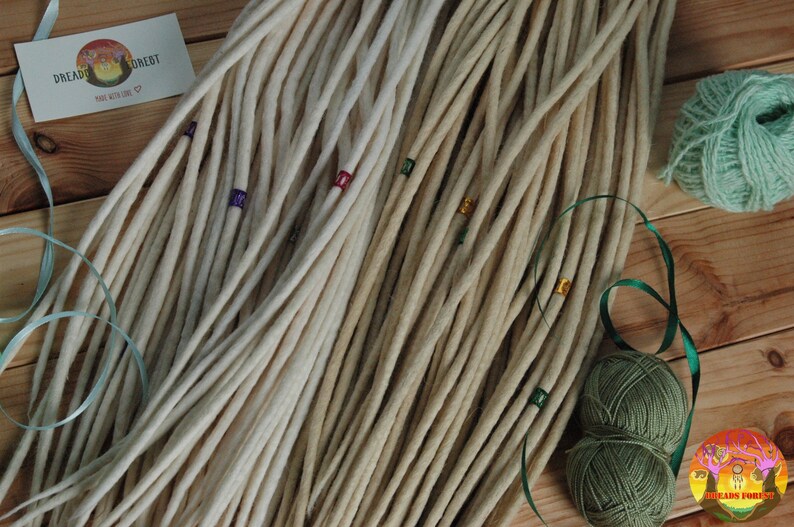 May include: A bundle of off-white wool dreadlocks with colorful beads. The dreadlocks are arranged in a fan shape on a wooden surface. There are two balls of yarn in the background, one green and one dark green.