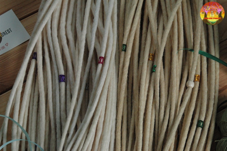 May include: Two sets of white synthetic dreadlocks with colorful beads. The dreadlocks are arranged in a bundle and are ready to be added to hair.