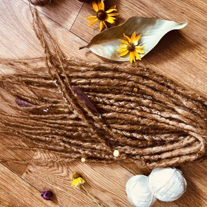 K&ouml;nnte beinhalten: Braune Dreadlocks mit dekorativen Perlen und einer Feder, angeordnet auf einer Holzoberfl&auml;che. Das Bild enth&auml;lt gelbe Blumen, ein Blatt und zwei Kn&auml;uel wei&szlig;e Wolle. Das Dread5 Forest-Logo ist unten links sichtbar.