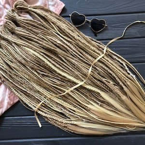 May include: A bundle of blonde and light brown synthetic dreadlocks, arranged on a dark wooden surface. The dreadlocks are tied together with a thin, light-colored ribbon. Heart-shaped sunglasses rest nearby. The Dreads Forest logo is visible.